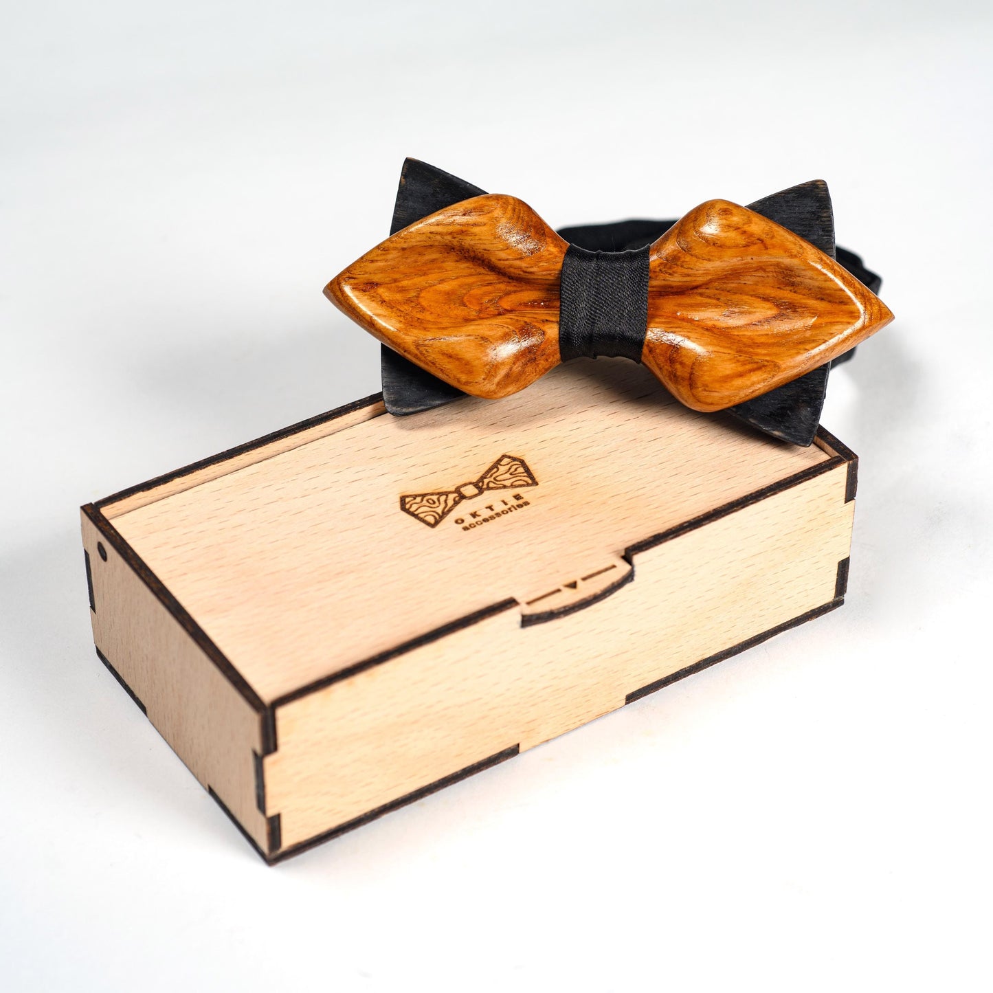 A wooden bow tie in a light brown color with a black bow tie backing. The bow tie is in a wooden box with the text "OXTIE WOODCRAFTING" engraved on the top.
